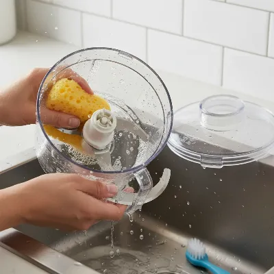 A person carefully cleaning the disassembled parts of a food processor (bowl, lid, blade) with soapy water and a sponge in a kitchen sink, emphasizing hygienic maintenance.