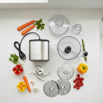 Food processor components laid out on a kitchen counter, showing the motor base, work bowl, lid, S-blade, and various discs for slicing and shredding, with fresh vegetables nearby.